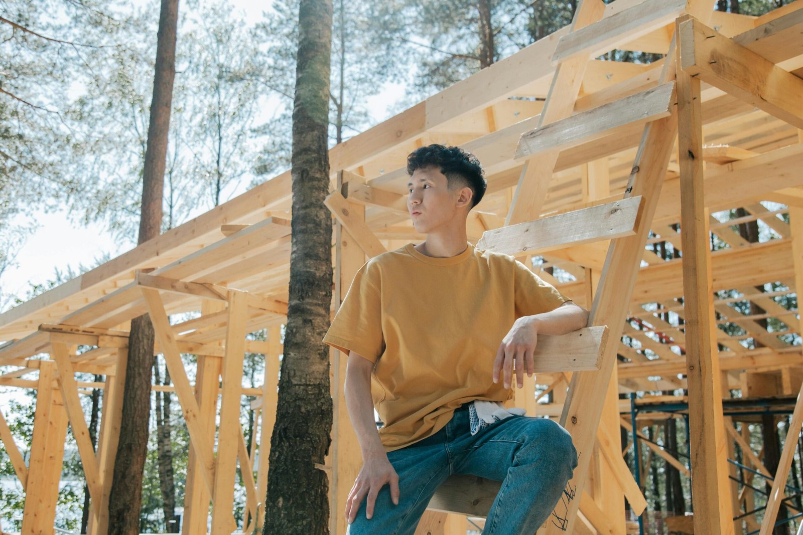 A young man sitting on a ladder at a forest construction site surrounded by wooden beams.